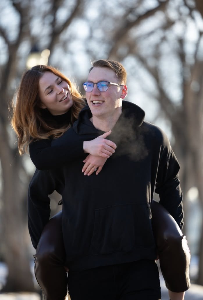A woman on her partner's back, both laughing, in a bare winter woodland